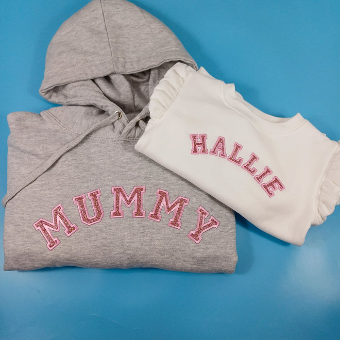 A gray hoodie with the text 'MUMMY' printed in pink on the front, along with a pink hood, presented next to a white, long-sleeved baby top with the name 'Hallie' printed in pink.