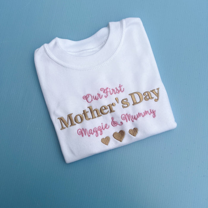 A white baby onesie with 'Our First Mother's Day' printed in gold and pink text, along with personalized names 'Maggie & Mummy' and heart shapes.
