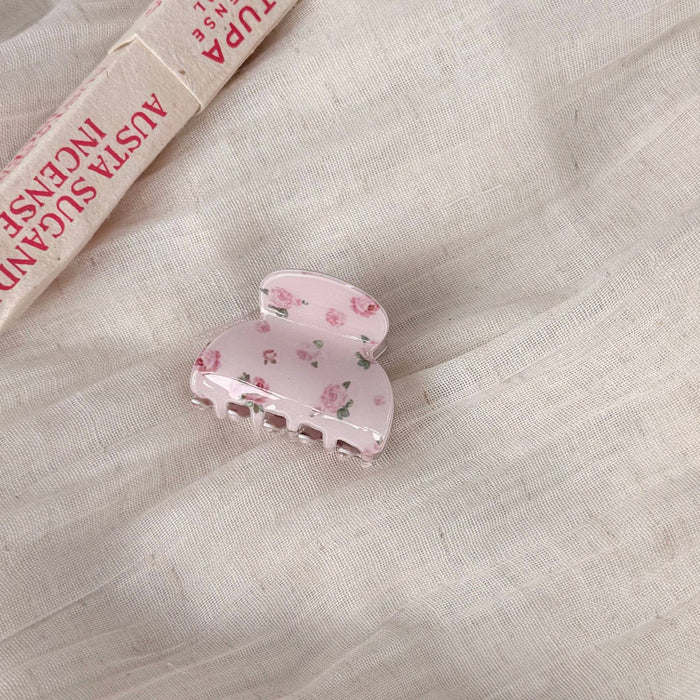 A light pink hair claw clip with a delicate rose pattern, resting on a textured beige linen fabric