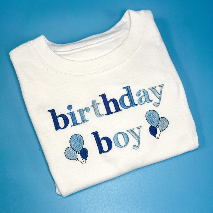 A folded white T-shirt with the text 'birthday boy' and balloon designs embroidered on the front.