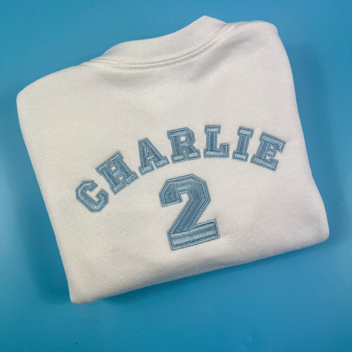 A white sweatshirt with the name 'Charlie' and the age '2' printed in a blue varsity style font on the front and back.