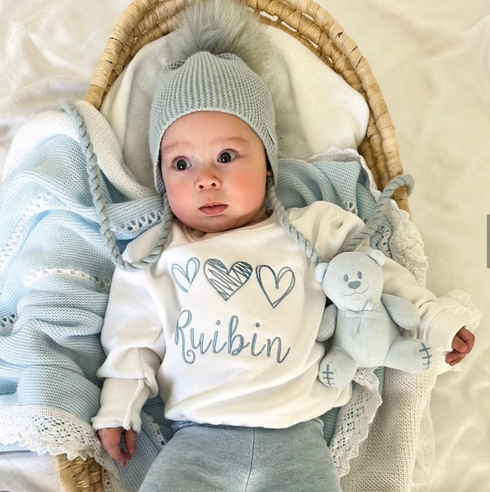 Baby in a basket wearing a white shirt with 'Ruibin' and heart designs, surrounded by soft fabrics.