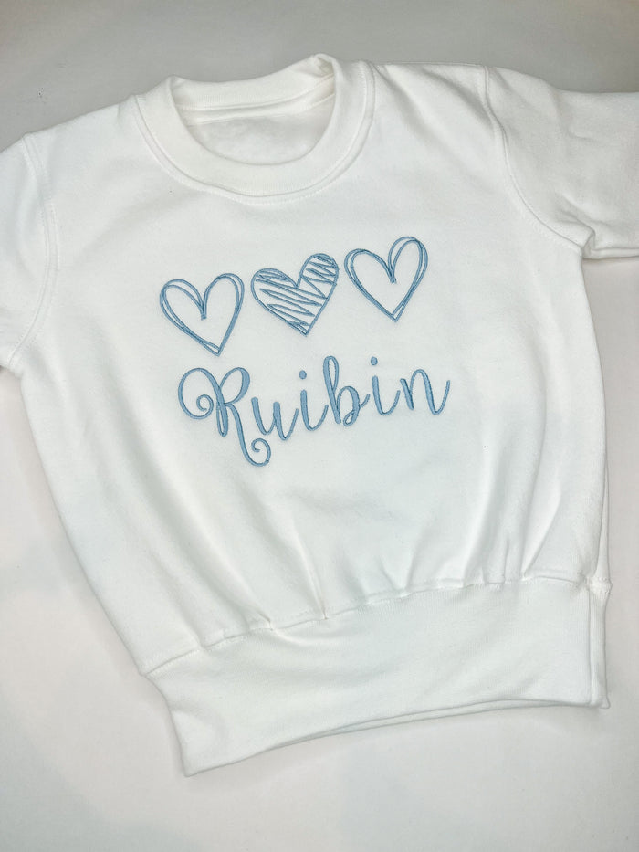 A white sweatshirt with fleece lining, featuring three blue scribble style hearts and the name 'Ruibin' in blue cursive text.