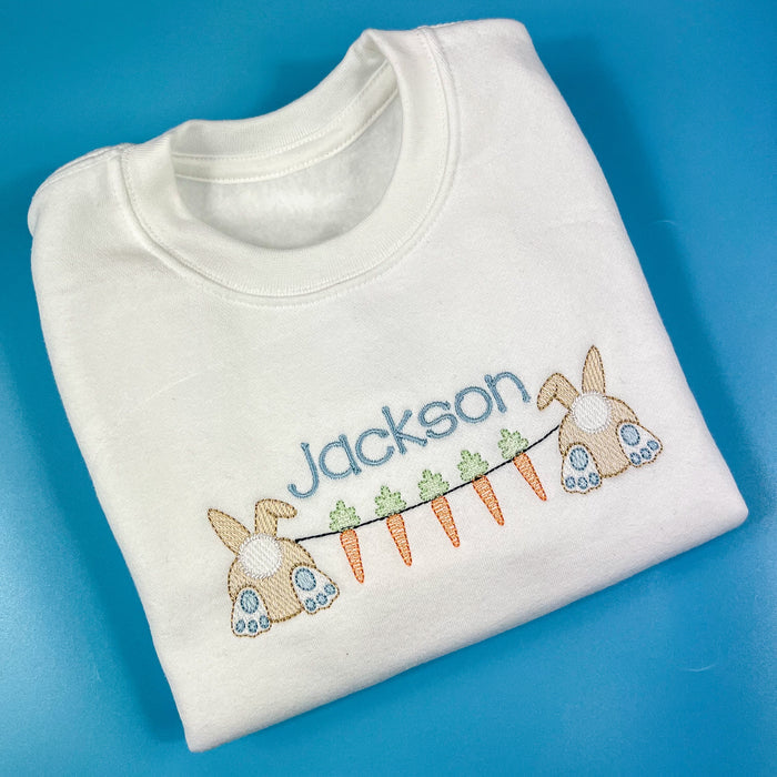 Folded white sweatshirt with embroidered design of bunnies and carrots on a blue background
