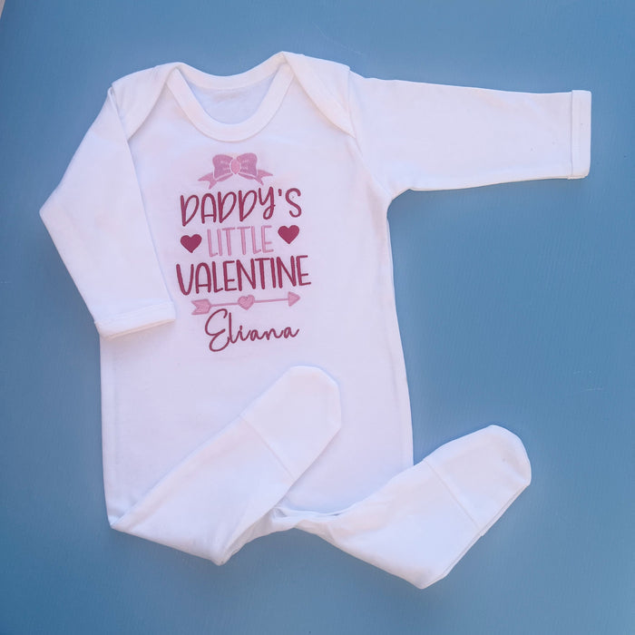 A white romper with 'Daddy's little valentine' printed on the front, along with a small heart and the name 'Elana'. The romper is for a child and has a pair of white booties attached.