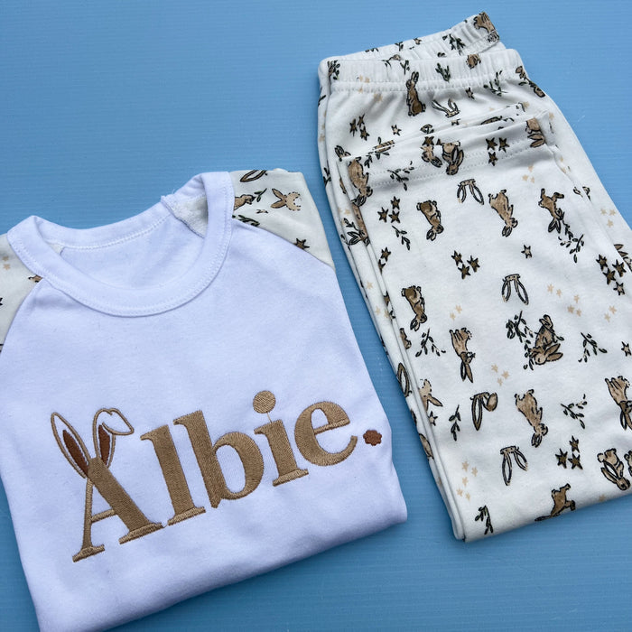 Children's Easter-themed pyjamas laid out on a flat surface, with the top piece displaying the name 'Albie' in a decorative font and the bottom piece adorned with a floral and rabbit pattern.