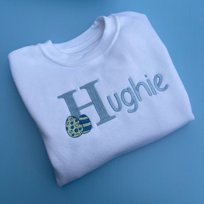 White jumper with the name Hughie embroidered on the front with an Easter egg
