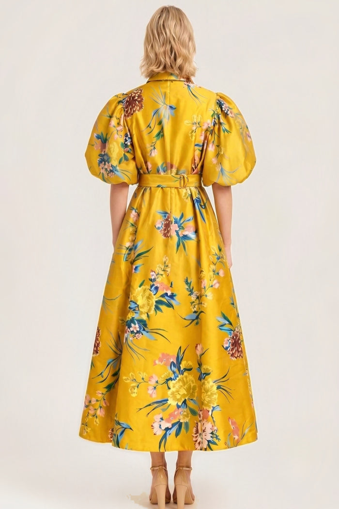 A woman viewed from the back wearing a bright yellow midi dress with a colorful floral print, puff sleeves, and a matching belt, set against a plain beige studio background.