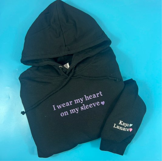 A black hoodie with the text 'I wear my heart on my sleeve' on the front and a name 'Kaya' with a heart symbol below it on the left sleeve. The hoodie is displayed on a blue background.