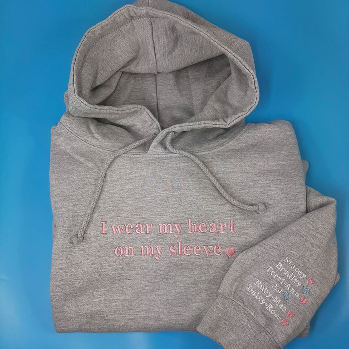 A grey hoodie with the text 'I wear my heart on my sleeve' on the front and names with a heart symbol below it on the left sleeve. The hoodie is displayed on a blue background.