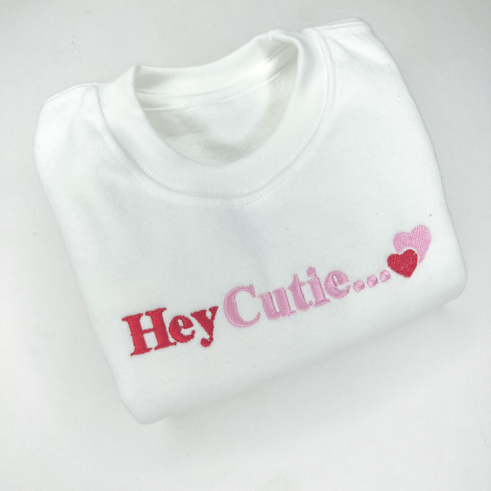 White sweatshirt with hey cutie embroidered on the front in pink and red