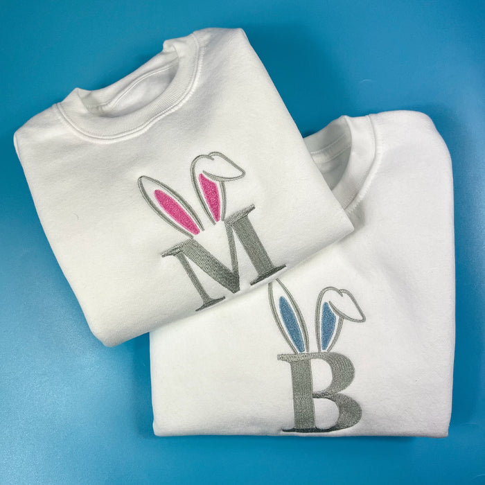 Two white sweatshirts with embroidered bunny ears designs and letters 'M' and 'B' on the front.