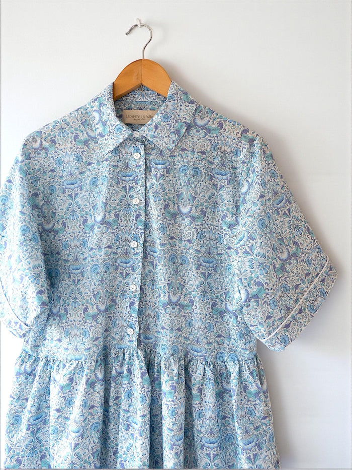 May include: A light blue floral print dress with short sleeves and a ruffled hem, hanging on a wooden hanger. The dress features a button-down front and white trim on the sleeves. The fabric has a delicate pattern of blue and purple flowers.