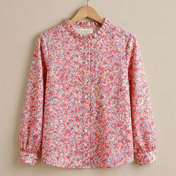 May include: A long-sleeved pink floral blouse with a ruffled collar and button-down front. The blouse features a delicate pattern of small pink and purple flowers and green leaves. The shirt is hanging on a wooden hanger.