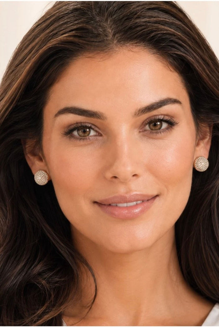 A close-up portrait of a woman with dark brown hair wearing textured, round, silver-toned stud earrings against a soft beige background.