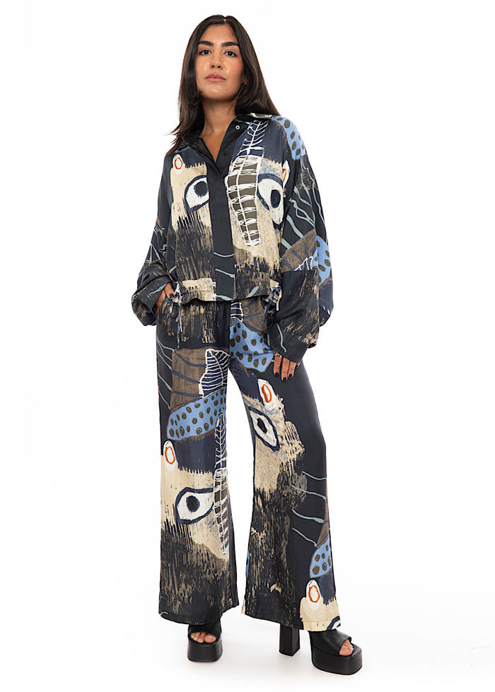 A model wearing wide leg Silk trousers with multicoloured pattern throughout. Styled with matching silk shirt with bat wing sleeves and black high heels.
