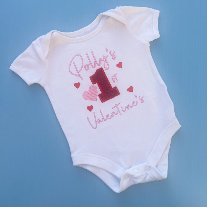 White baby vest with Valentine's Day design including the text 'Polly's 1st Valentine's Day' and decorative hearts.