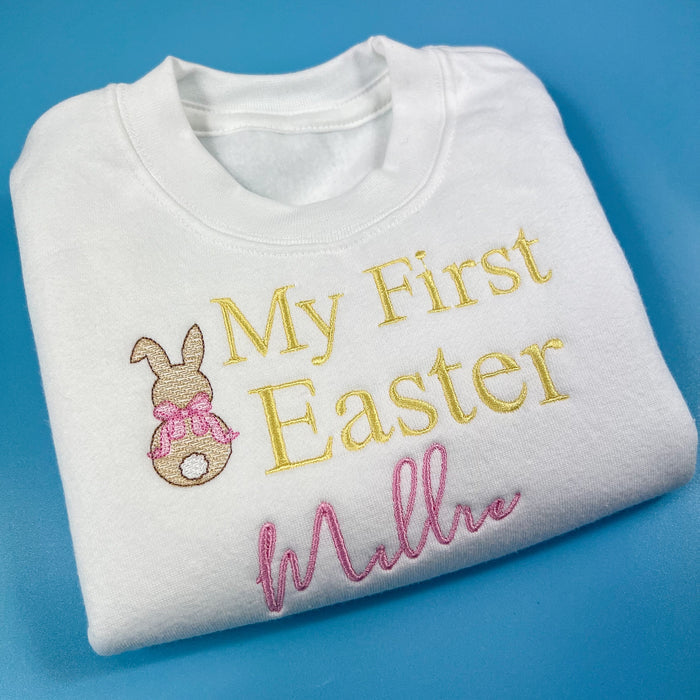 A white Easter-themed jumper for babies with the text 'My First Easter' and a decorative element, folded and displayed against a blue background.