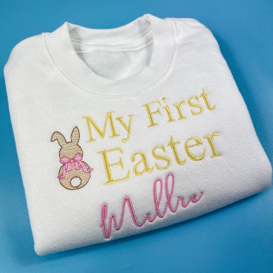A white Easter-themed jumper for babies with the text 'My First Easter' and a decorative element, folded and displayed against a blue background.