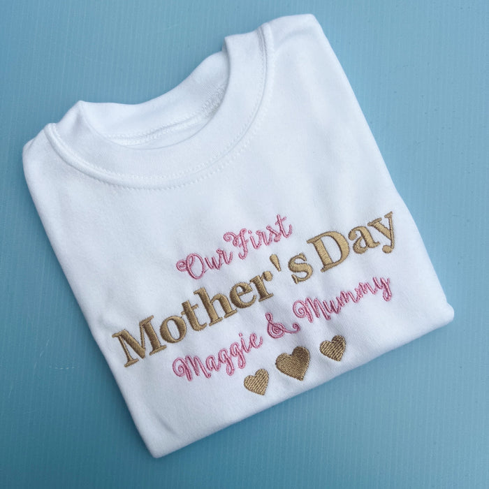 A white baby onesie with 'Our First Mother's Day' printed in gold and pink text, along with personalized names 'Maggie & Mummy' and heart shapes.