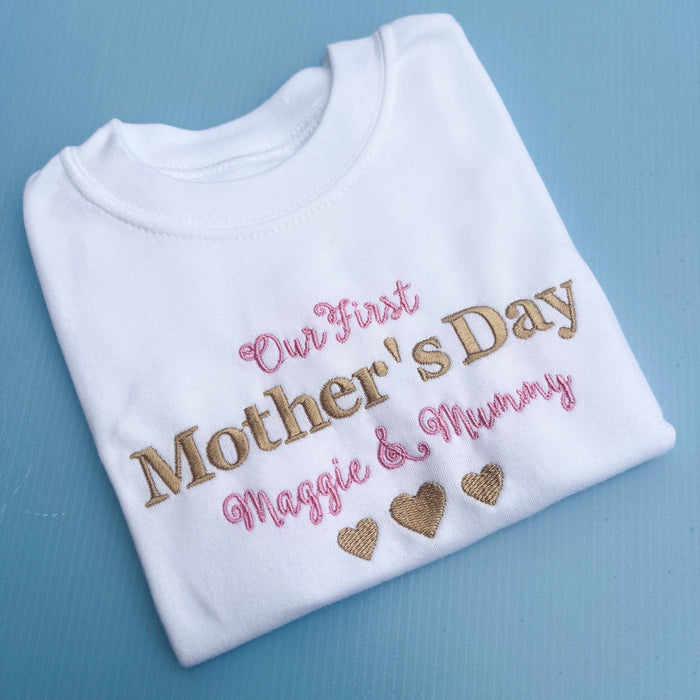 A white baby onesie with 'Our First Mother's Day' printed in gold and pink text, along with personalized names 'Maggie & Mummy' and heart shapes.
