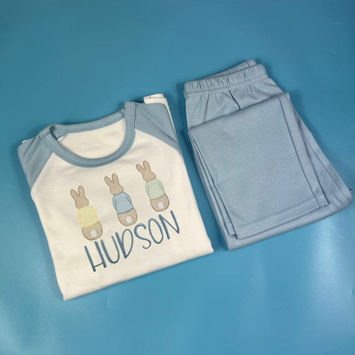 A set of children's pyjamas with a white t-shirt featuring bunny illustrations and the name 'Hudson' on it, along with blue pants.
