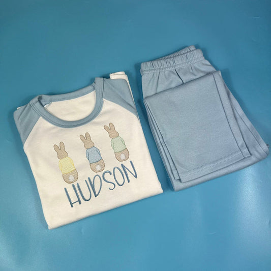A set of children's pyjamas with a white t-shirt featuring bunny illustrations and the name 'Hudson' on it, along with blue pants.