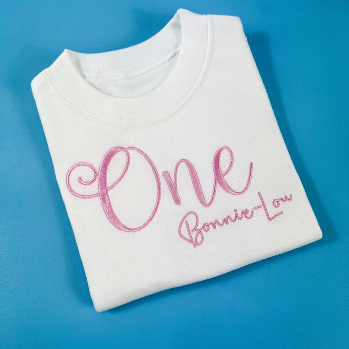 A white t-shirt with the number 'One' and the name 'Bonnie-Lou' embroidered in pink on the front.